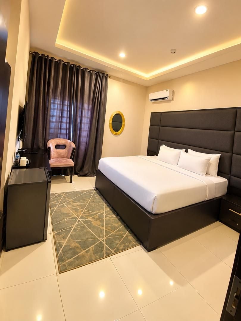 A hotel room with a bed, a desk, a chair, a mirror, and an air conditioning unit.