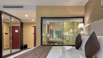 A hotel room with a large bed, a glass partition wall, and a view of the hotel's interior.