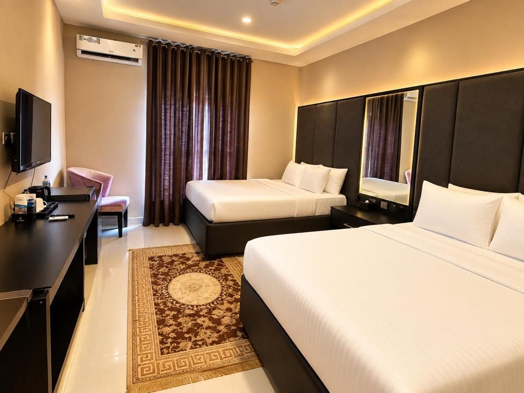 A hotel room with two beds, a desk, a television, and an air conditioning unit.