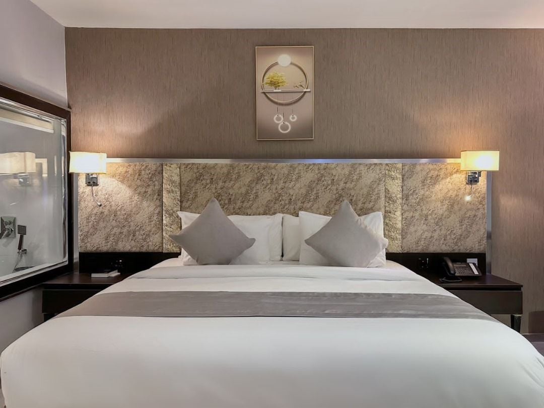 A hotel room with a large bed, two bedside lamps, a headboard with a circular light fixture, and a wall-mounted artwork.