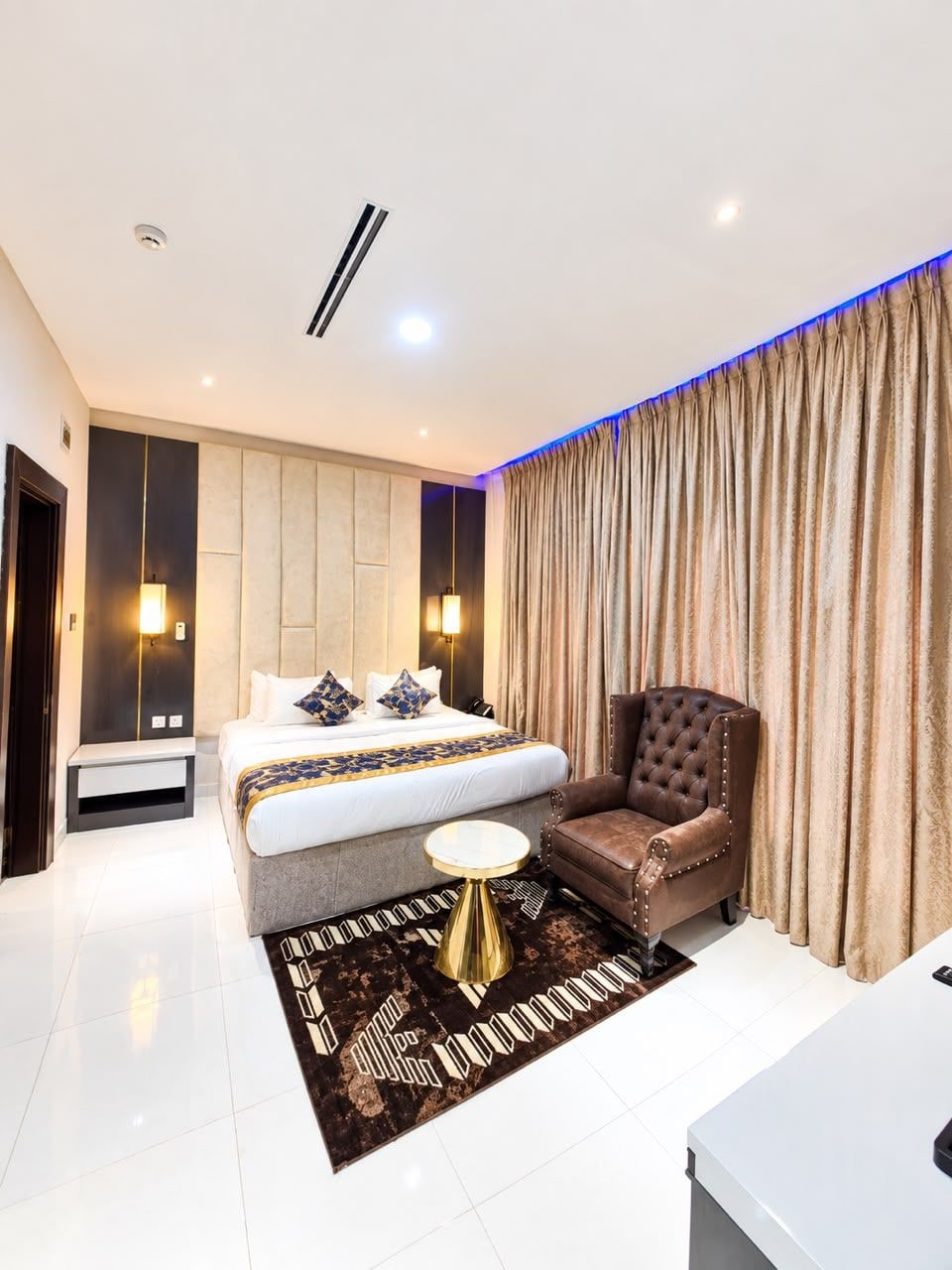 A modern hotel room with a large bed, a sitting area with a chair, a bedside table, and a small round table.