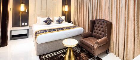 A modern hotel room with a large bed, a sitting area with a chair, a bedside table, and a small round table.