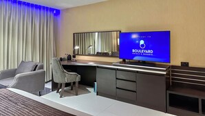 Una habitación de hotel moderna con un televisor de pantalla plana grande que muestra el nombre del hotel, un escritorio con una silla, un sofá y una ventana grande con cortinas.