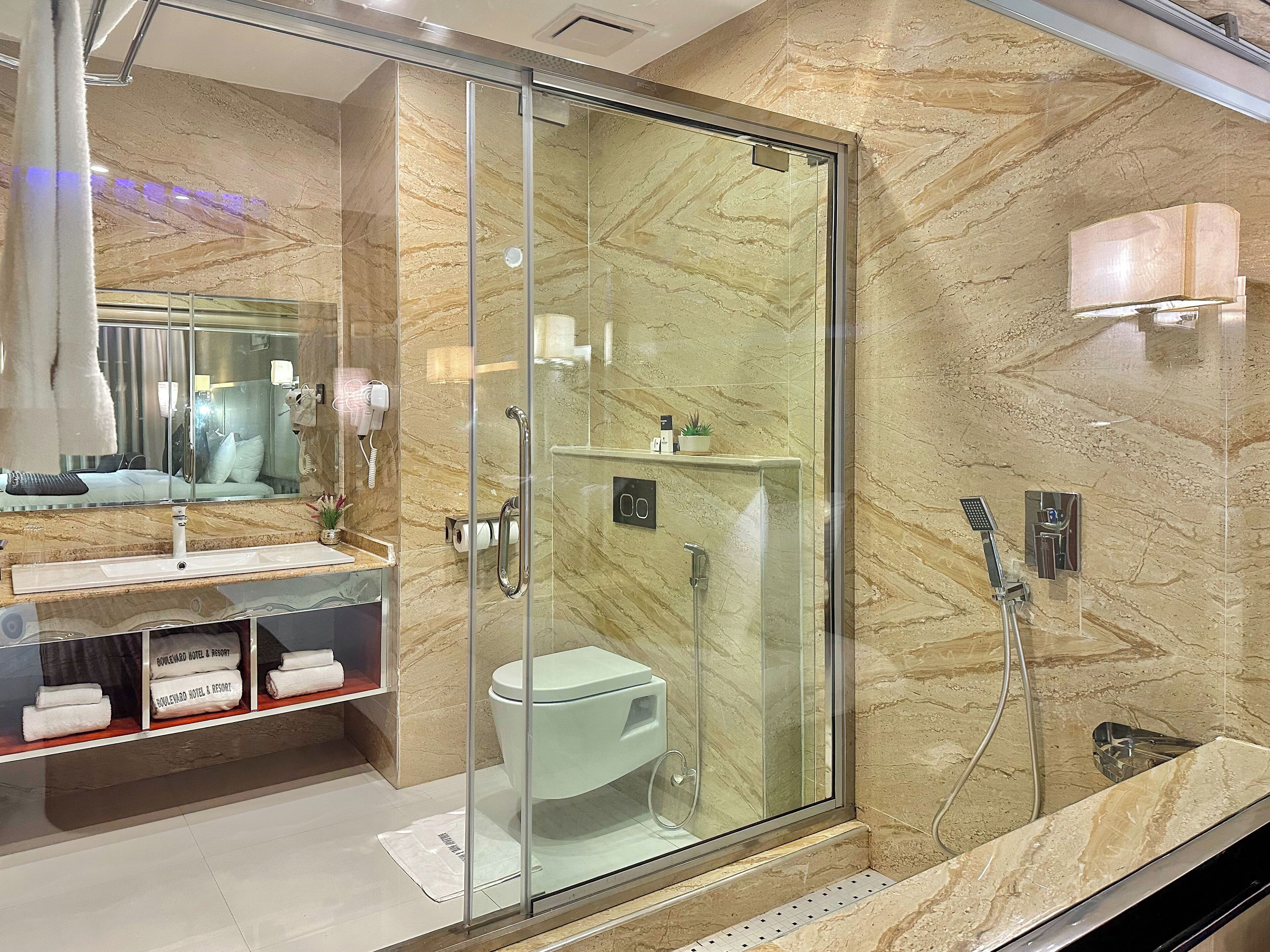 Un baño moderno con ducha envuelta en vidrio, pared con azulejos de mármol, tocador con espejo y estante con toallas.