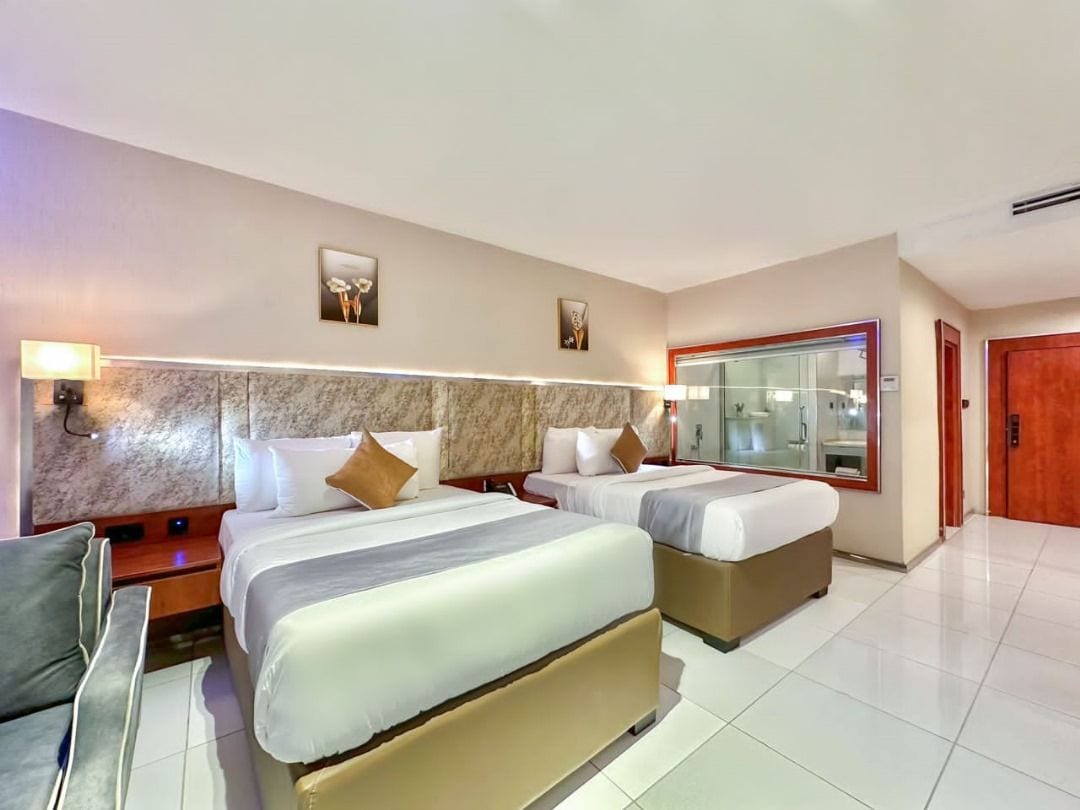 Habitación de hotel con dos camas, un sofá, un espejo grande y vista al exterior.