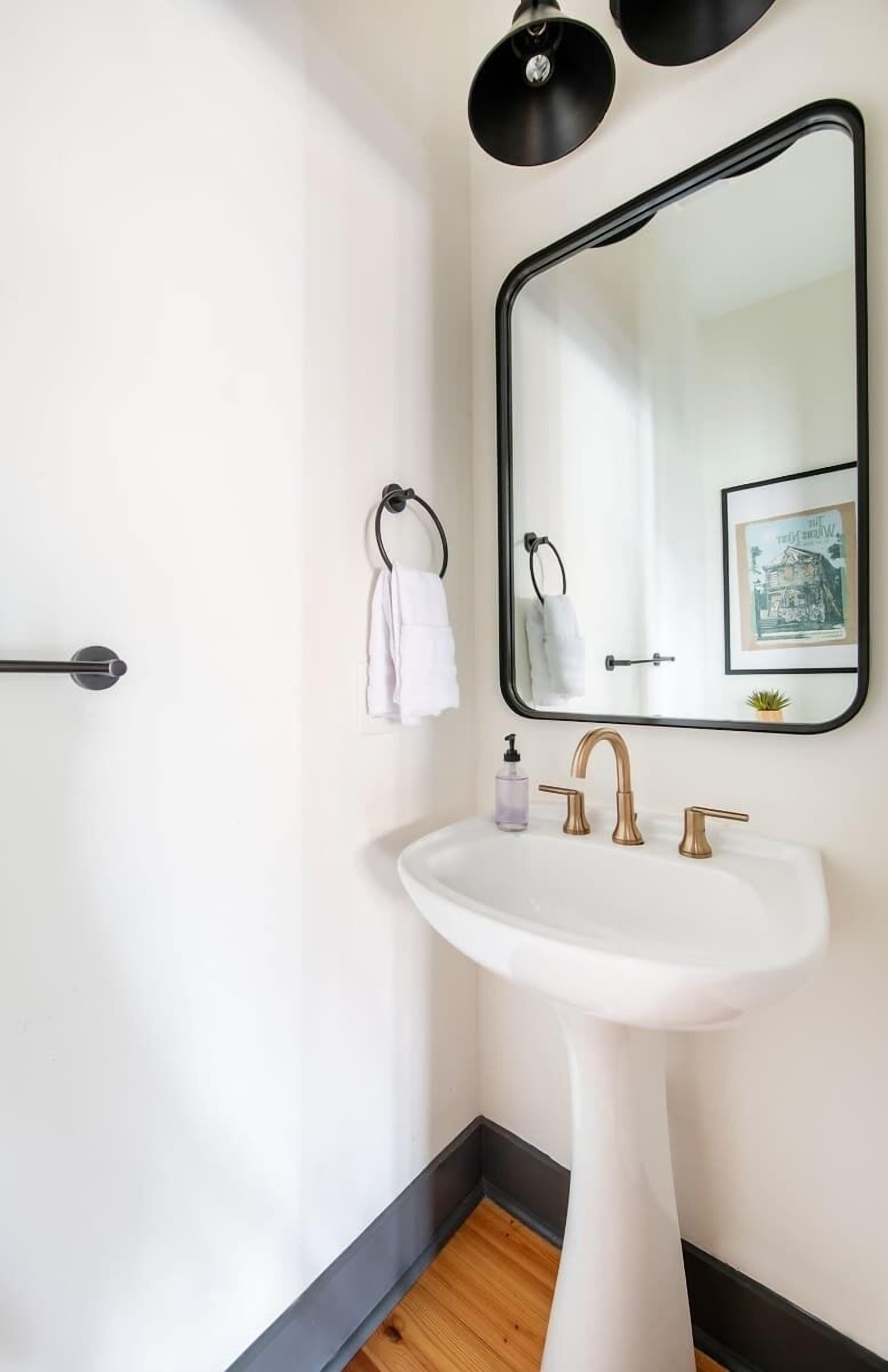 Une salle de bain avec un lavabo blanc, deux robinets dorés, un miroir à cadre noir, un porte-serviettes avec une serviette blanche, un tableau encadré et un plancher en bois.