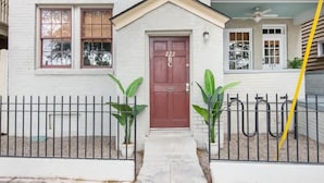 Una casa de dos pisos con puerta roja, fachada blanca y un balcón con plantas.