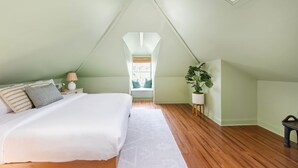 Un dormitorio con techo inclinado, cama con ropa de cama blanca, mesita de noche con lámpara y planta en maceta.