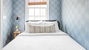 Un dormitorio con cama, una ventana con persianas, una lámpara de noche y una mesita con un florero.