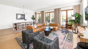 A spacious living room with a brown leather sofa, black patterned ottomans, a dining area with wooden furniture, and large windows with curtains. - Newly Built, 4bd Luxury Suite steps to King! (Charleston)