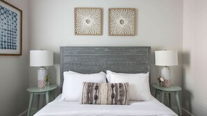 Una cama bien arreglada con sábanas blancas y dos almohadones decorativos, flanqueada por dos mesitas de noche con lámparas y jarrones. Encima de la cama, hay dos paneles decorativos en la pared.