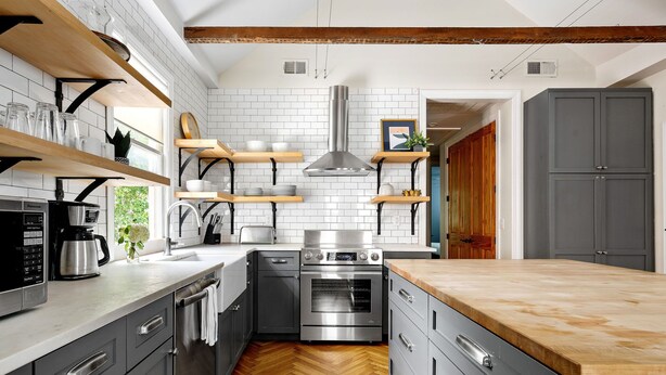 A modern kitchen with grey cabinetry, wooden shelves, a stainless steel range, and a herringbone wood floor.