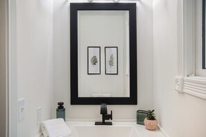 Un baño con un espejo grande, un lavabo blanco, una toalla, un dispensador de jabón y una pequeña planta en maceta.