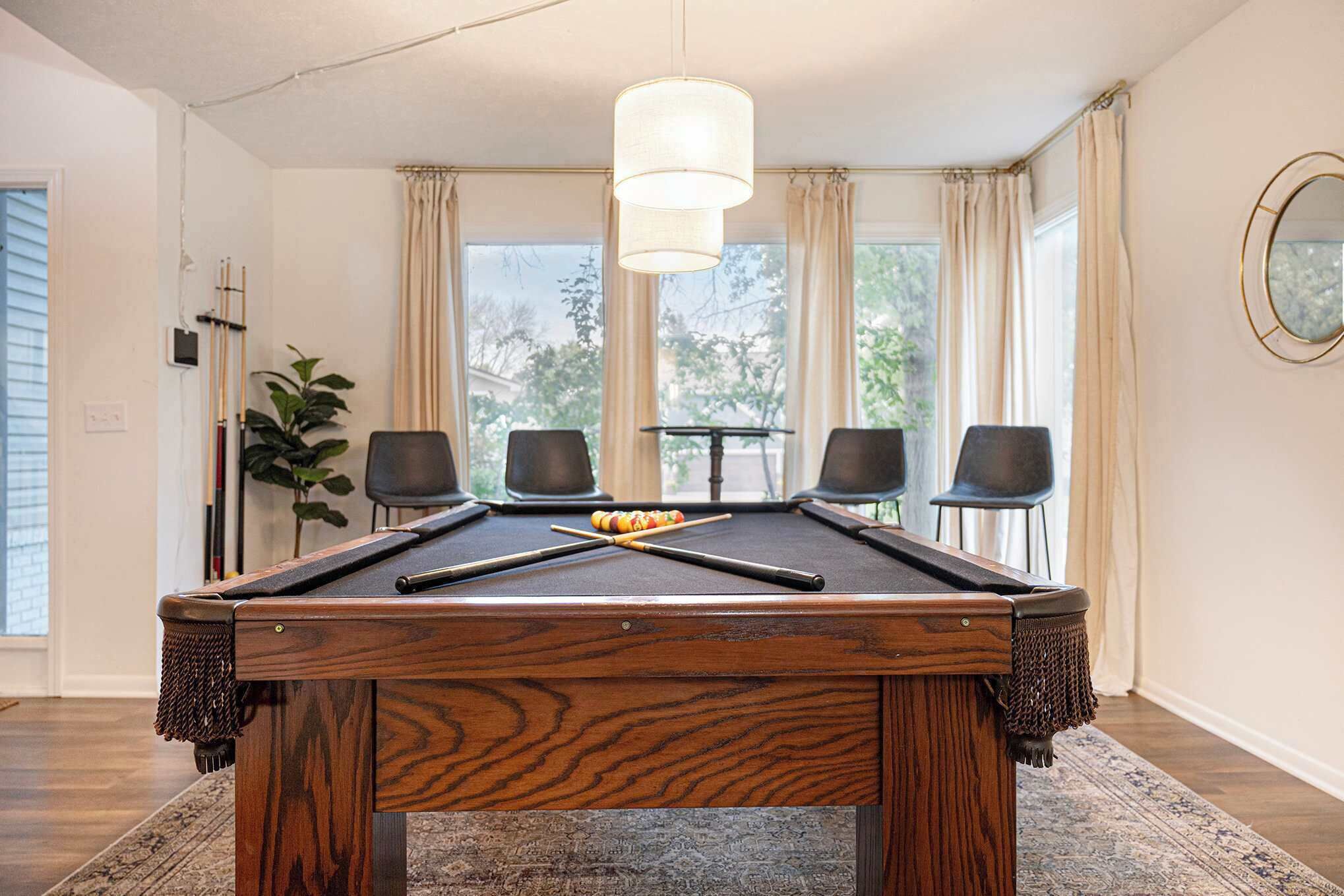 Una mesa de billar con taco y bolas, rodeada de cuatro sillas, en una habitación con ventanas amplias y una lámpara colgante.