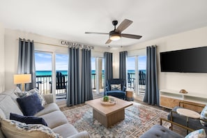 A living room with a view of the ocean, a ceiling fan, a sofa, a coffee table, and a TV on the wall.
