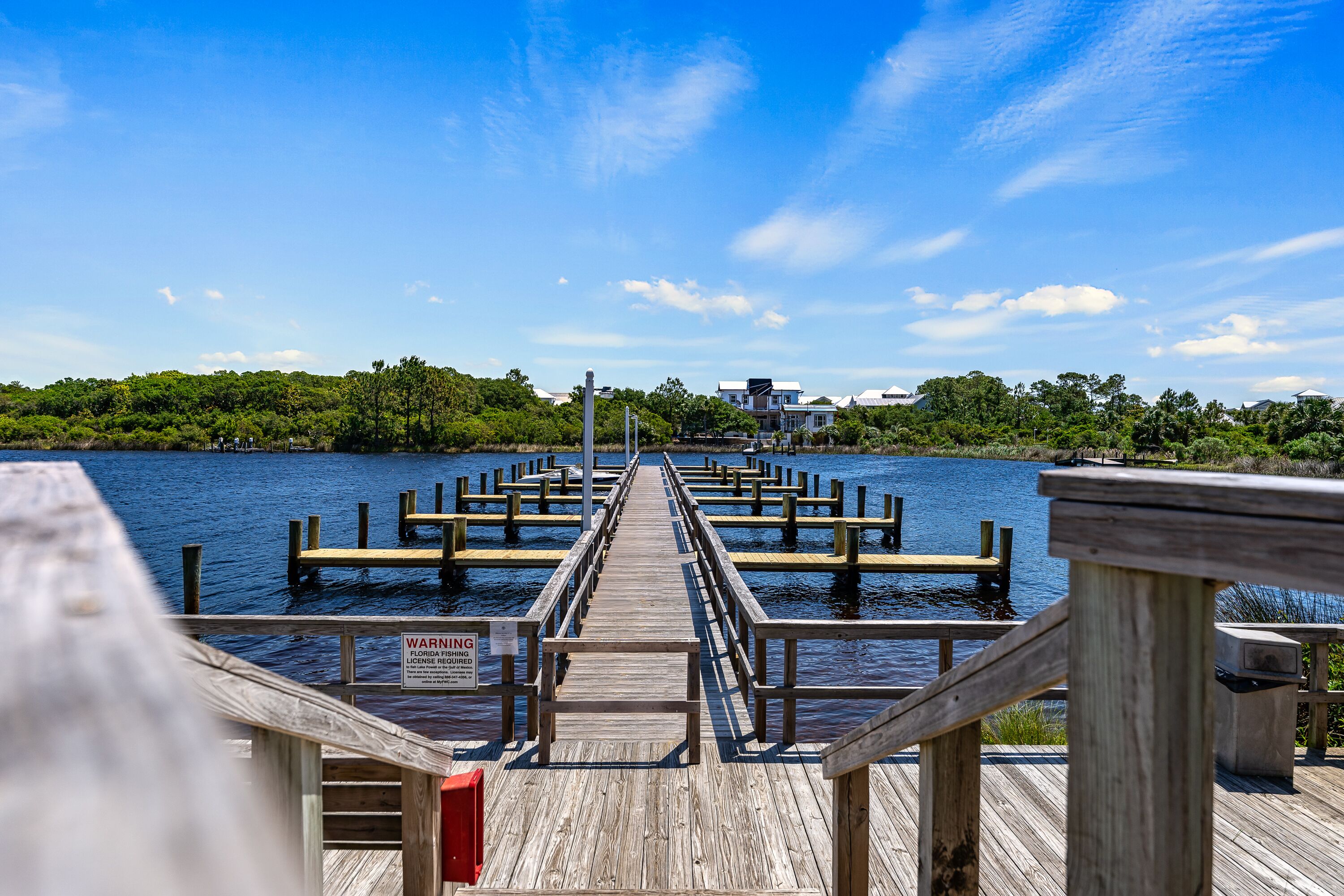 A wooden pier extending into a body of water with a warning sign, surrounded by greenery and buildings in the distance.