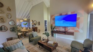Amplia sala de estar con una gran televisión de pantalla plana montada en una pared con diseño geométrico, comedor con mesa y sillas de madera, y una zona de estar acogedora con sofás y una mesa de centro.
