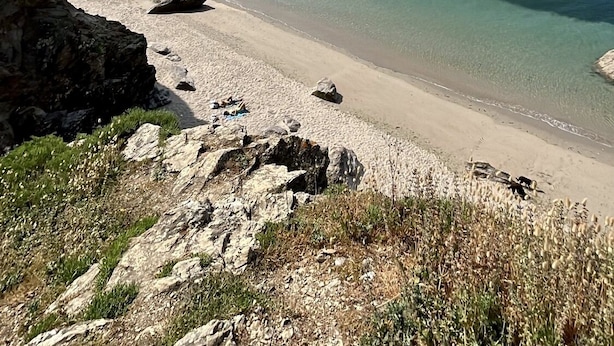 Une plage côtière aux eaux cristallines, avec un rivage rocheux et un voilier à l’horizon.