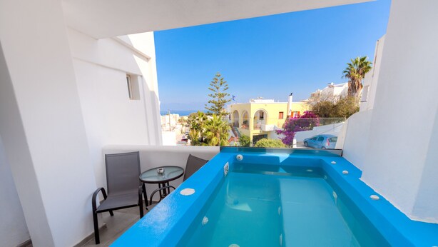 A rooftop pool with a blue edge, a small table, and a chair, overlooking a residential area with buildings and trees. - The Muses of Santorini (Santorini)
