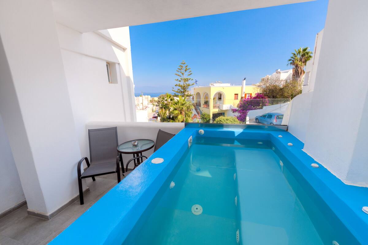 A rooftop pool with a blue edge, a small table, and a chair, overlooking a residential area with buildings and trees.