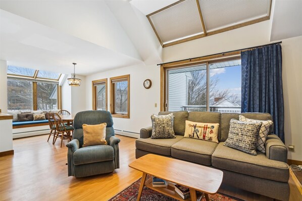 A spacious living room with a high ceiling, large windows, a sofa set, a dining area with a table and chairs, and a wooden coffee table. - Ski-in/ski-out 3 bedroom condo with pool and hot tub (Killington)