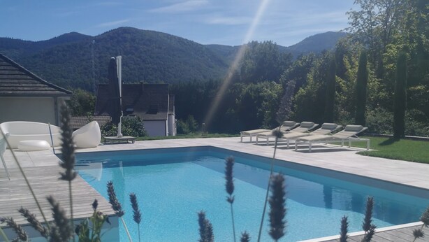 A rectangular swimming pool with a wooden deck, surrounded by greenery and mountains in the background.