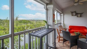 Ein Balkon mit Blick auf einen Wald, ein rotes Sofa, Korbstühle und ein Deckenventilator.