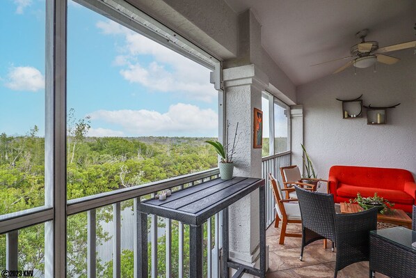 A balcony with a view of a forest, a red sofa, wicker chairs, and a ceiling fan.