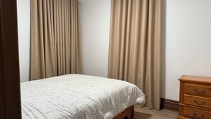 Ein Schlafzimmer mit einem Bett, beige farbenen Vorhängen, einer Holzkommode und einer weißen, strukturierten Tagesdecke.
