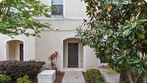 Un edificio con fachada color beige, puerta negra y un pequeño jardín con banco y plantas.