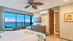 Ein Hotelzimmer mit einem großen Bett, einem Deckenventilator, Blick auf den Ozean, einer Holzkommode und einem gerahmten Bild an der Wand.