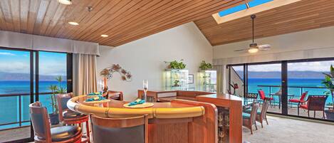 Un comedor moderno con techo de madera, ventanas amplias con vistas al océano y una barra con taburetes.
