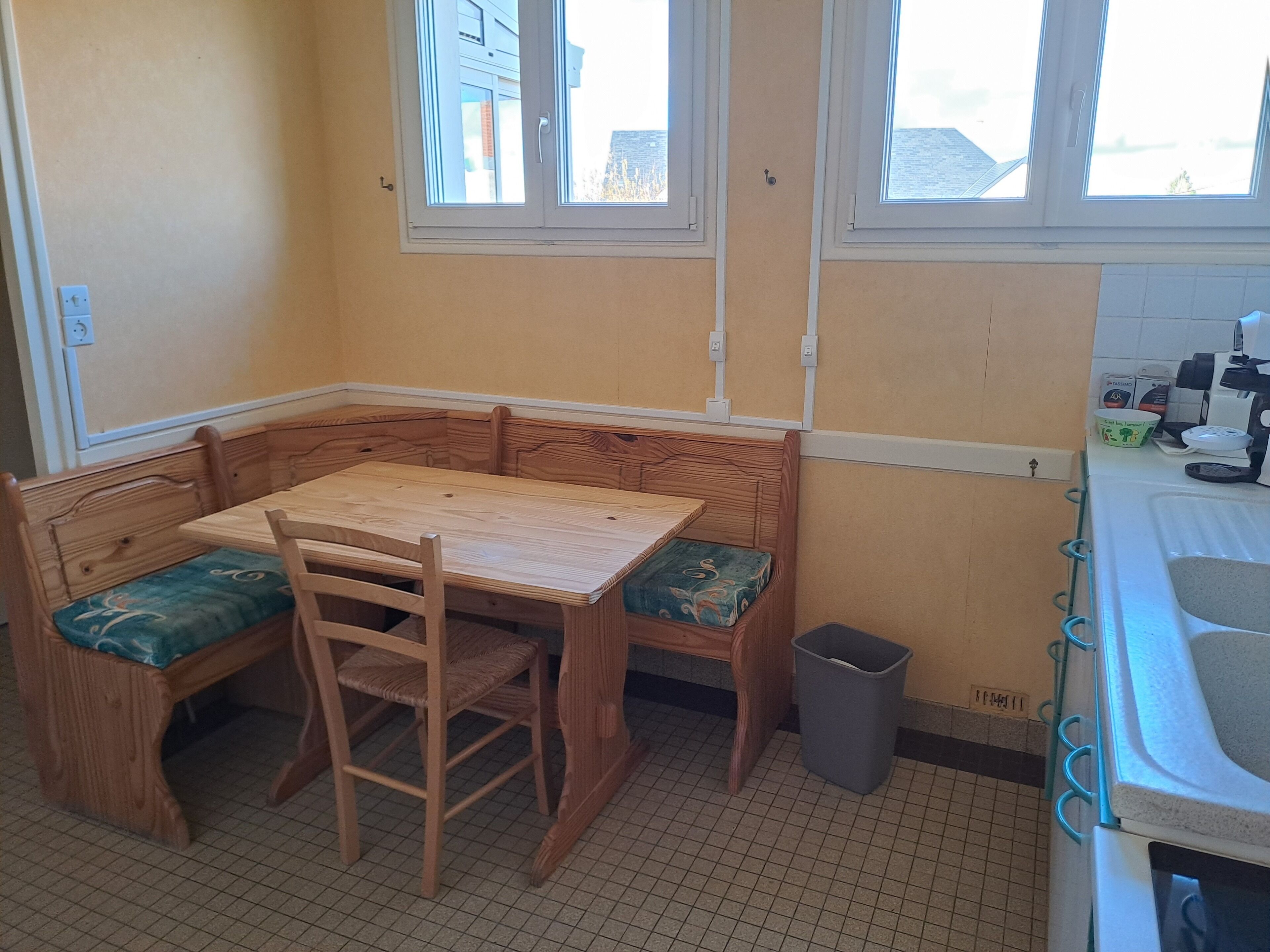 Cozinha no canto com mesa e banco de madeira, lixeira e janela com vista para prédios.
