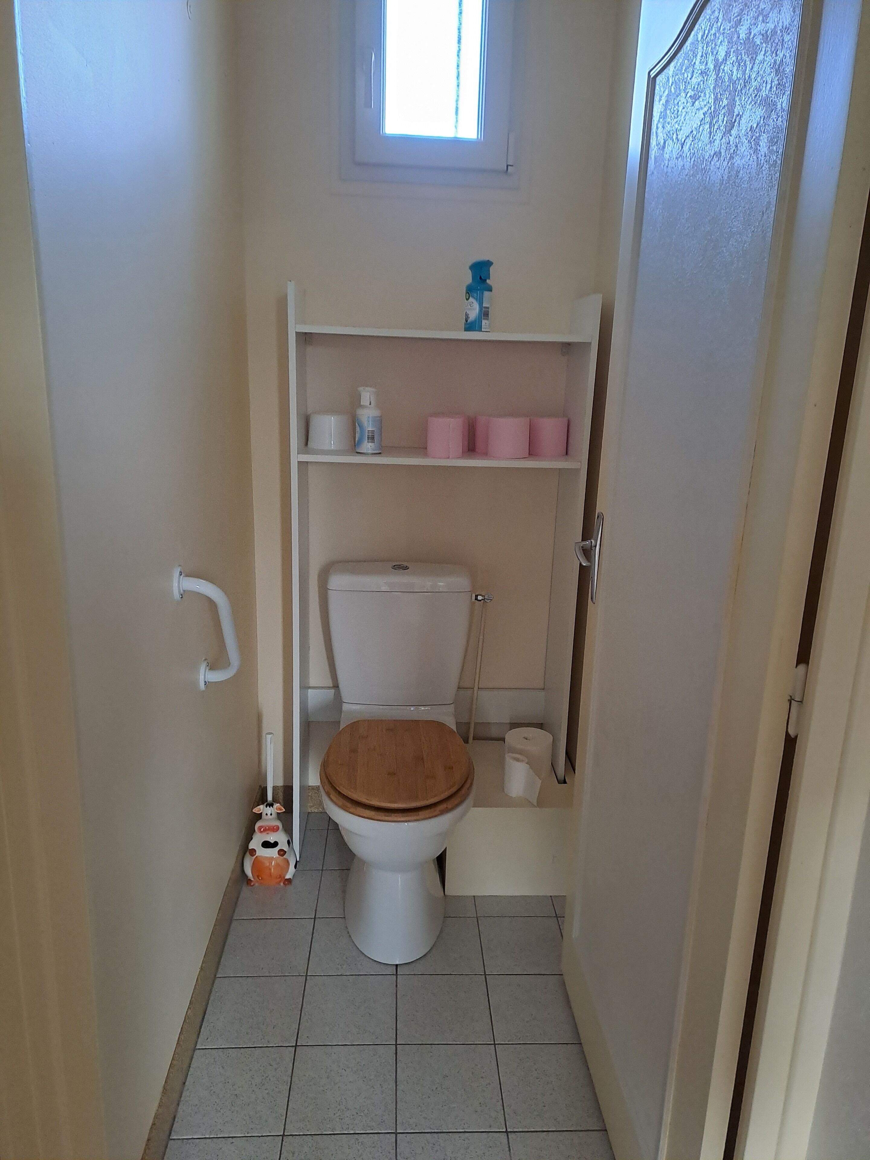 A small bathroom with a toilet, a shelf with toiletries, and a window above the toilet.