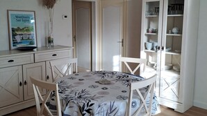 Un comedor con una mesa preparada para cuatro personas, rodeada de sillas blancas. A la derecha, un armario blanco con puertas de cristal alberga vajilla y vasos. Sobre la mesa cuelga una lámpara colgante blanca.