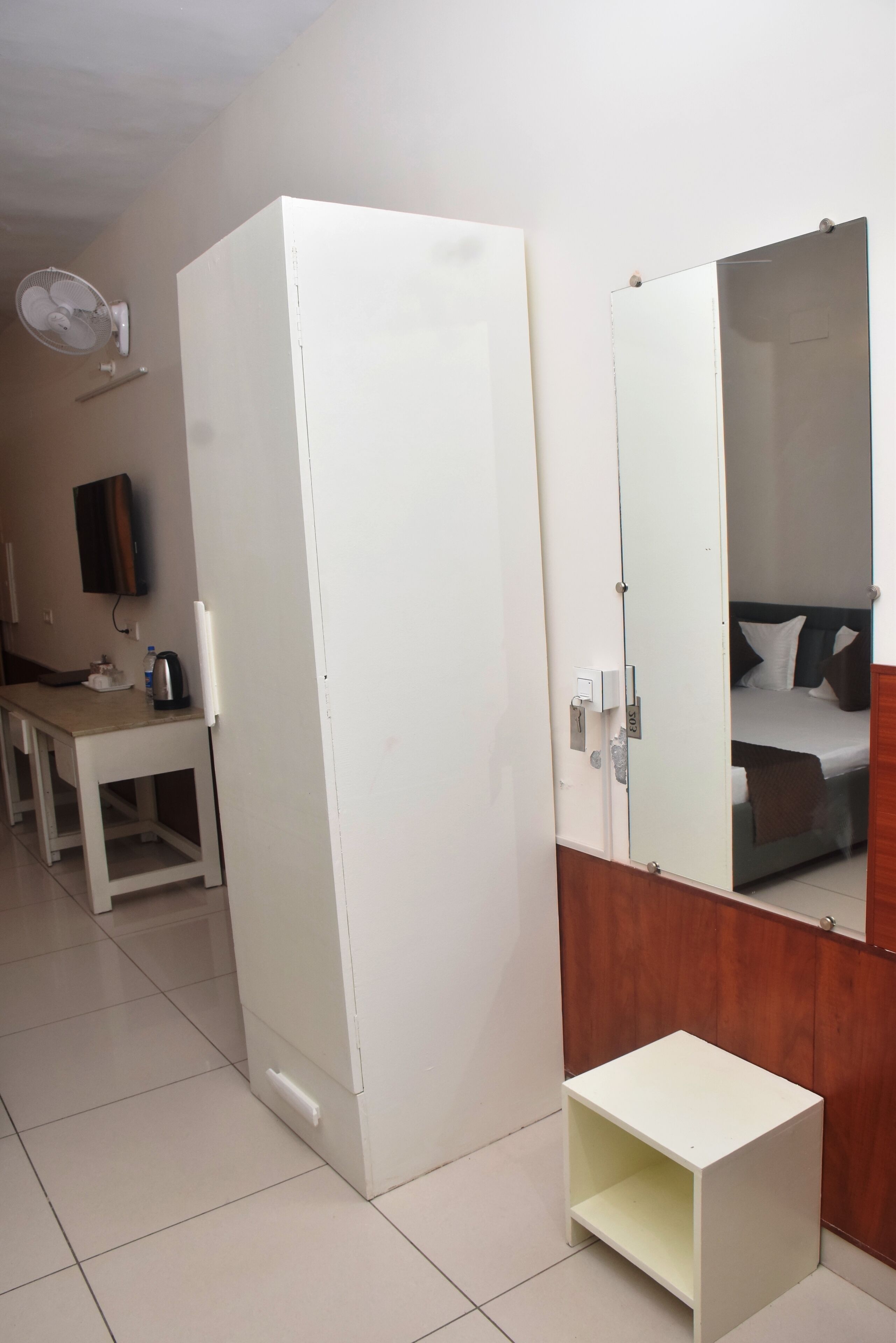 Une chambre d’hôtel comprenant un lit, une table de chevet, une petite armoire blanche, un miroir, une garde-robe blanche, un bureau avec une chaise, une bouilloire et un ventilateur.
