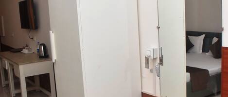 Une chambre d’hôtel comprenant un lit, une table de chevet, une petite armoire blanche, un miroir, une garde-robe blanche, un bureau avec une chaise, une bouilloire et un ventilateur.