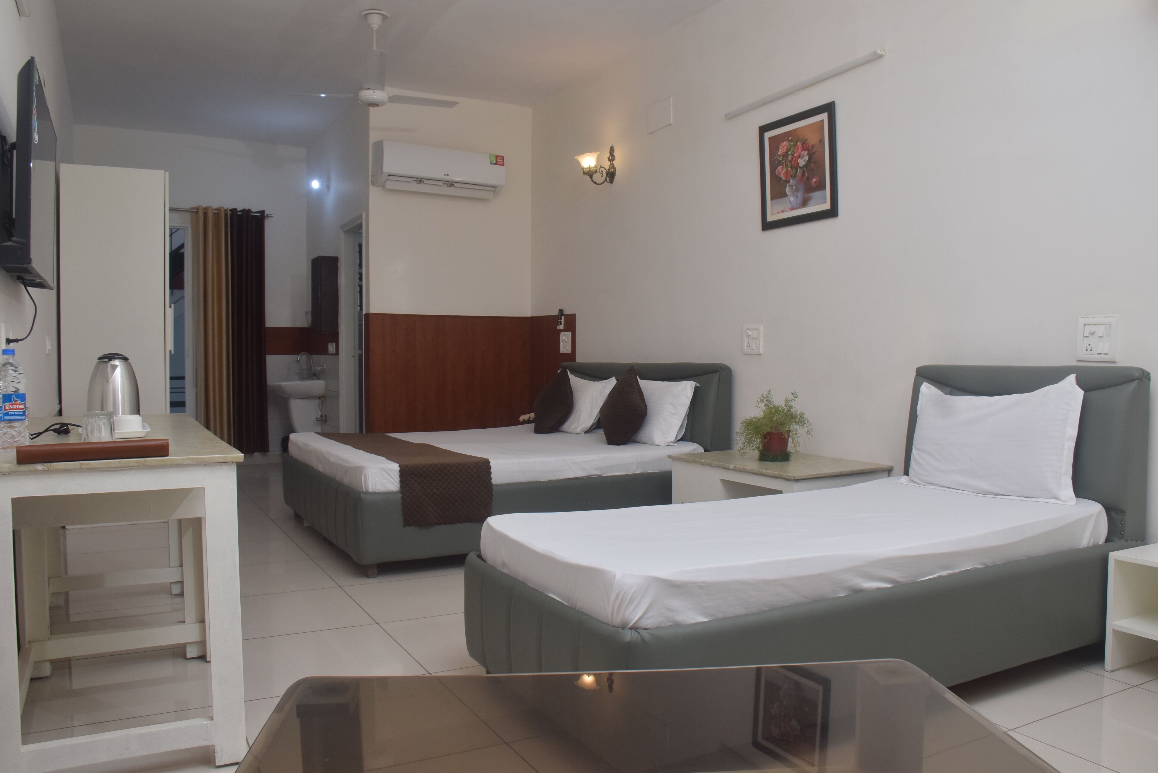 A hotel room with two beds, a desk, a chair, a small table, a potted plant, and a wall-mounted air conditioner.