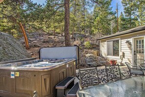 A hot tub in a backyard with a patio table and chairs, surrounded by trees and a house. - Red Rocks Retreat ~ Hot Tub, Game Room, Views, Privacy (Morrison)