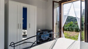Un dormitorio con cama, armario, escritorio con un sombrero y vistas a la vegetación a través de un ventanal.