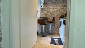 Un couloir étroit avec un plancher en bois, deux chaises marron, une petite table, un luminaire suspendu et un coin cuisine avec un mur en briques et des armoires blanches.
