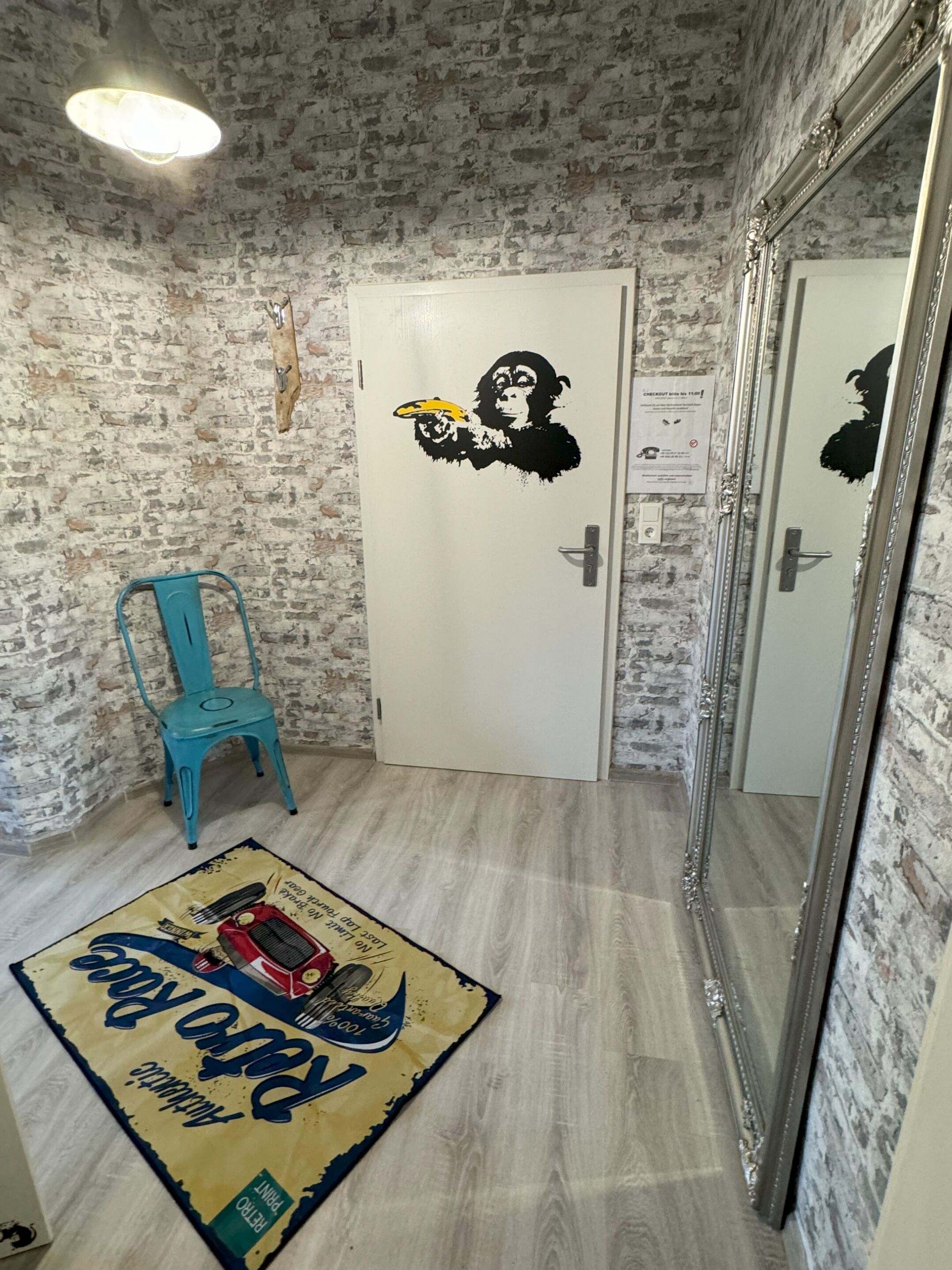 Ein Zimmer mit einer Ziegelwand, einer weißen Tür mit einem Affen-Sticker, einem blauen Stuhl und einem Teppich mit einem Vintage-Autodesign.