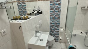 Une salle de bain avec un lavabo, des toilettes et une baignoire, tous de couleur blanche. Un miroir est placé au-dessus du lavabo, et on peut voir un porte-serviettes. Les murs sont recouverts de carreaux décorés.