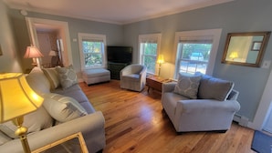 Ein Wohnzimmer mit einer Couch, einem Sessel und einem kleinen Tisch mit einer Lampe. Zwei Fenster mit Jalousien, ein Fernseher und ein Spiegel an der Wand sind ebenfalls vorhanden.