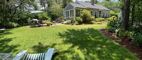 A well-maintained lawn with a white lounge chair, a house with a porch, and surrounding greenery.