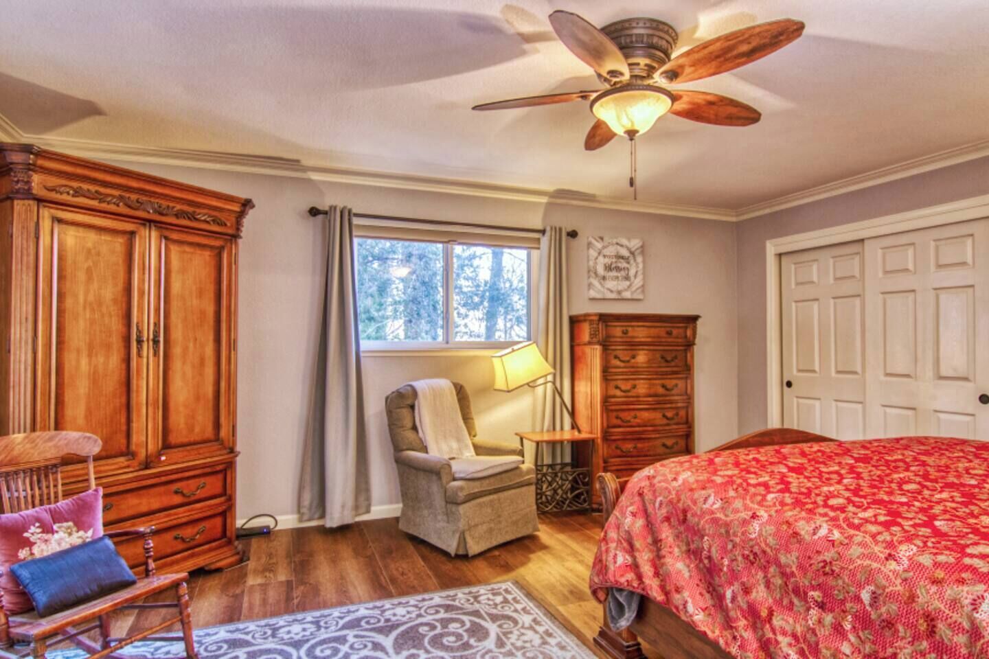 A bedroom with a wooden wardrobe, a chair, a bed with a floral bedspread, a ceiling fan, and a window with curtains.