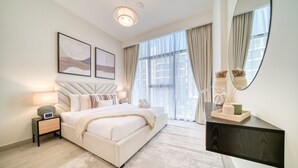 A modern bedroom with a large bed, bedside tables, a nightstand, a mirror, and a view of the outside through large windows. - Ravishing 2BR at Azizi Riviera 30, Al Merkadh by Deluxe Holiday Homes (Dubai)