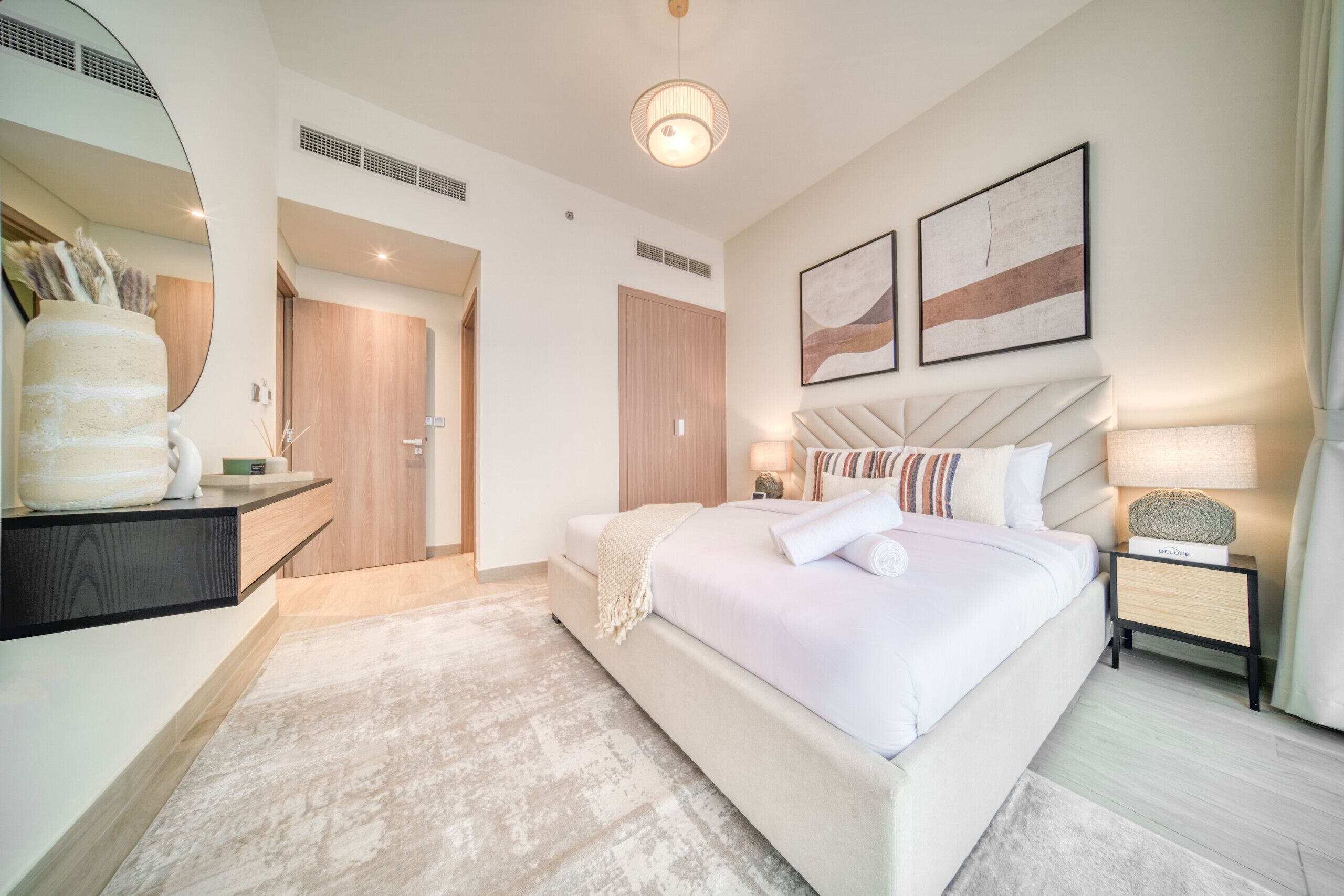 A modern hotel room with a large bed, a bedside table with a lamp, a mirror, and artwork on the wall.