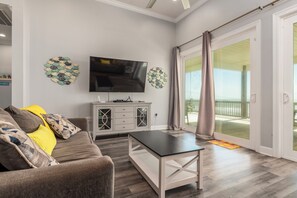 Una sala de estar moderna con un televisor de pantalla plana, un sofá gris con cojines amarillos y estampados, un mueble blanco con cajones y un vidrio que da a un balcón.