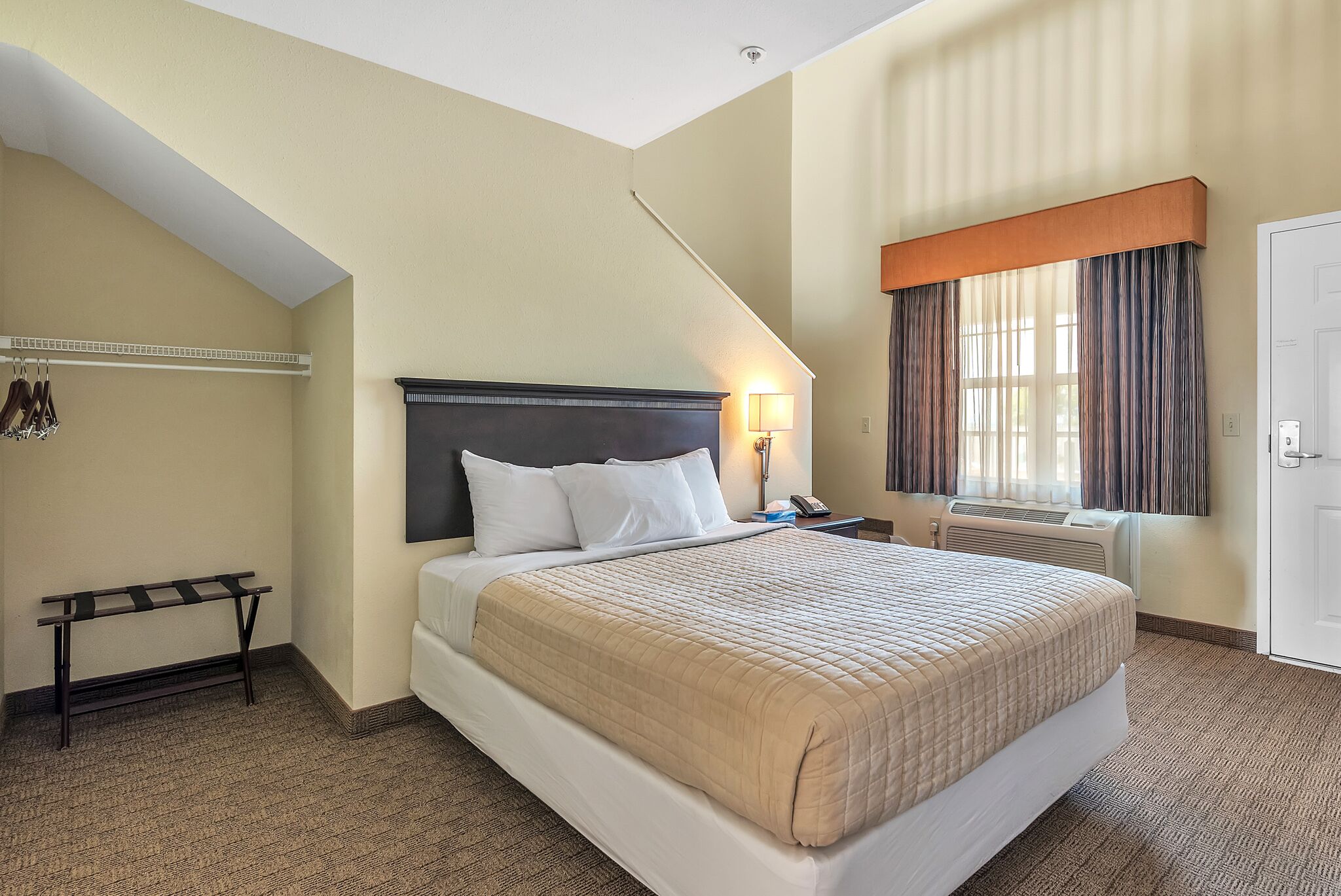 A hotel room with a large bed, a bedside table, a lamp, a window with curtains, and a coat rack.